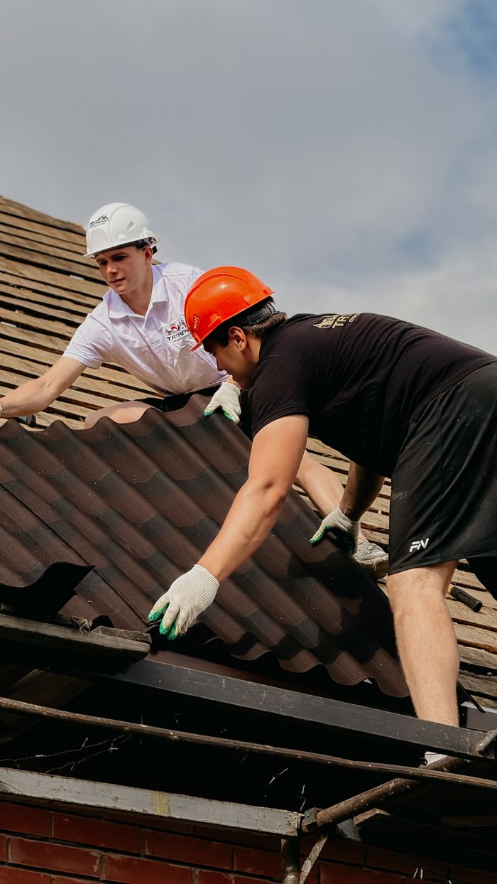 About Two construction workers install roofing wearing safety helmets on a sunny day.
