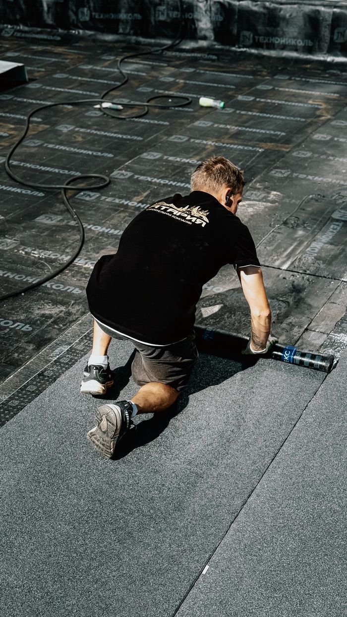 A construction worker installs asphalt roofing material under the sun, demonstrating professional craftsmanship.